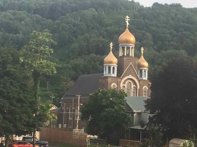 An church with bright copper domes and double Patriarchal crosses sits against a forested backdrop