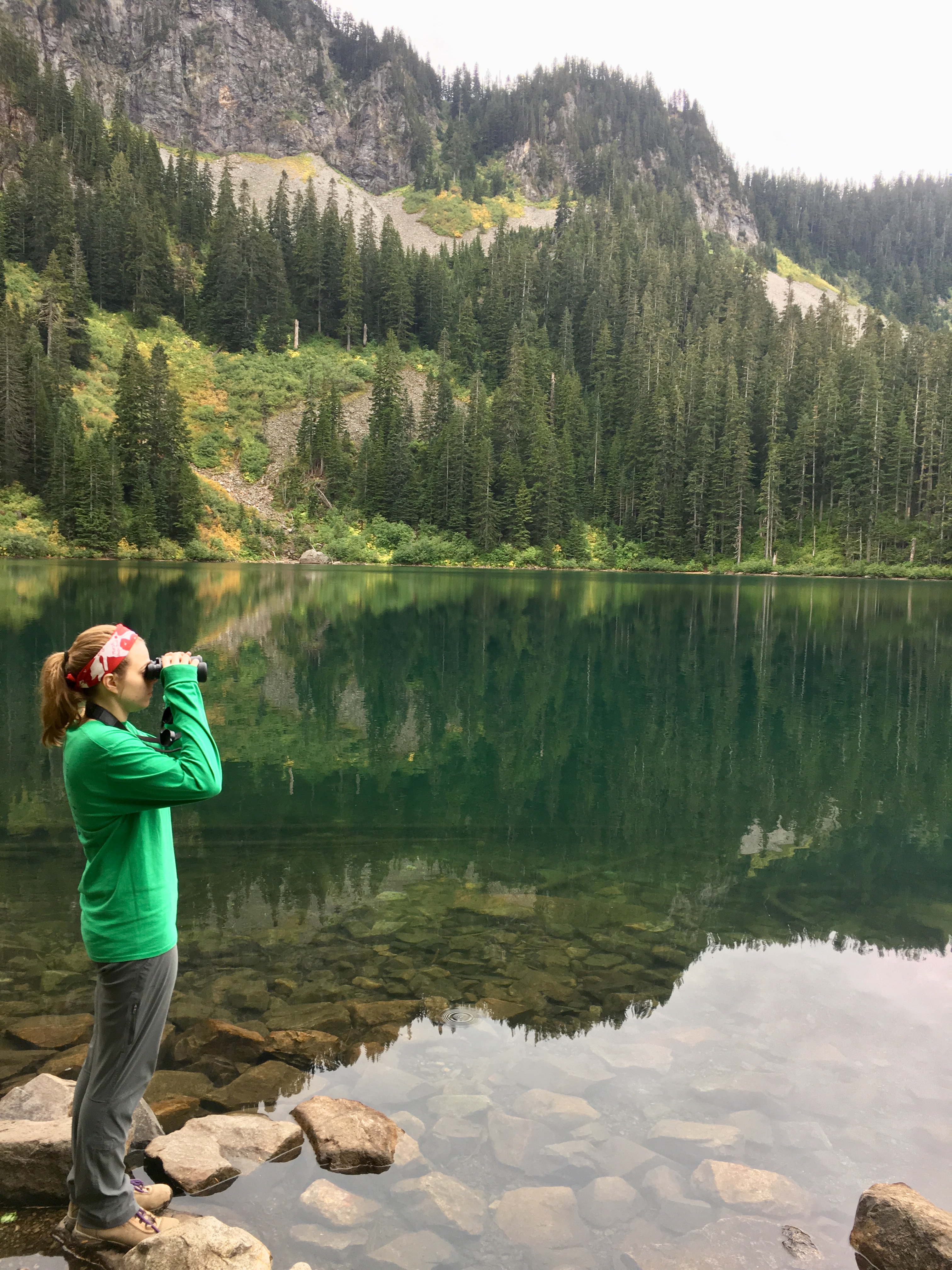 A young woman in hiking clothes holds up binoculars along a mountain lakeshore.