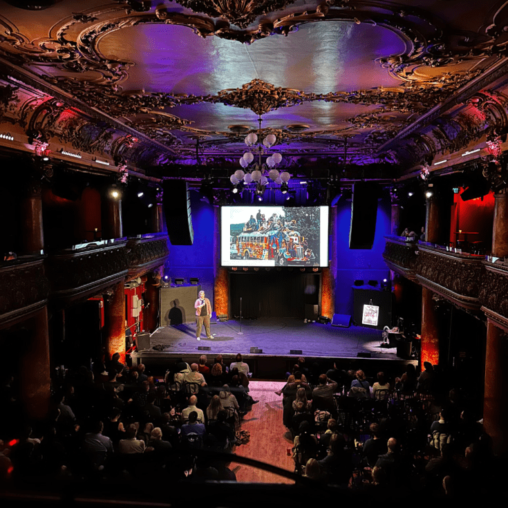 Scene of a person speaking on a stage to a full crowd in a venue with an old, decorative ceiling