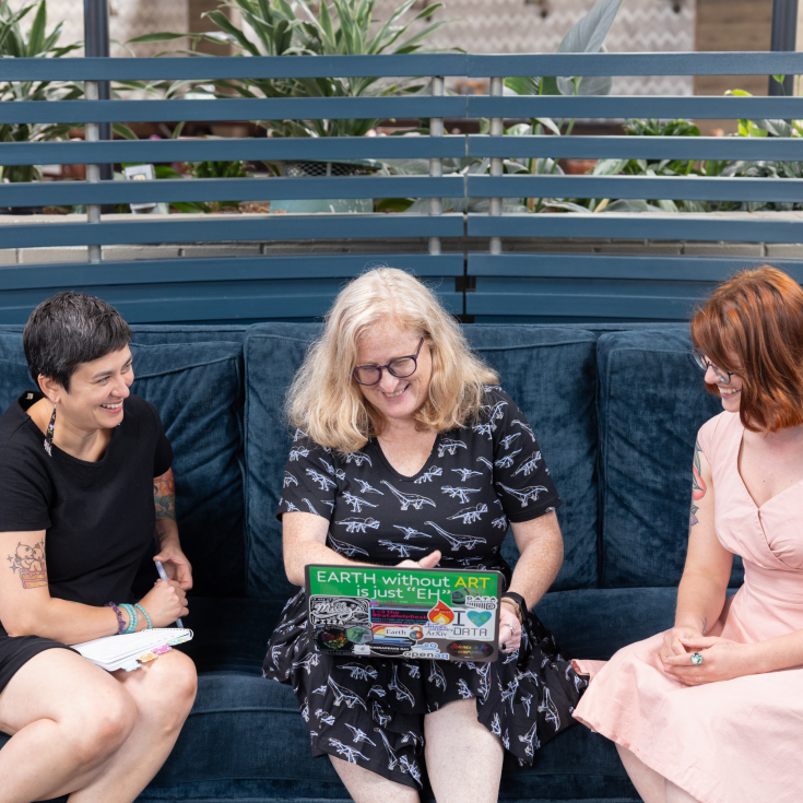 three people sit on a couch, one is holding a laptop with a bumbersticker that reads "Earth without art is just 'eh'"